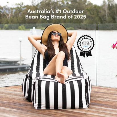 Woman relaxing on a black and white striped bean bag by a waterfront, with a brand badge claiming 'Australia's #1 Outdoor Bean Bag Brand of 2025'.