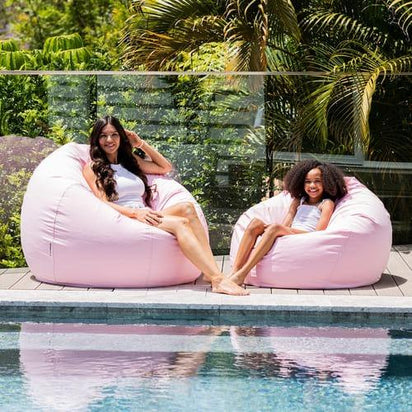 Two women sitting on pink bean bags by a pool with greenery in the background